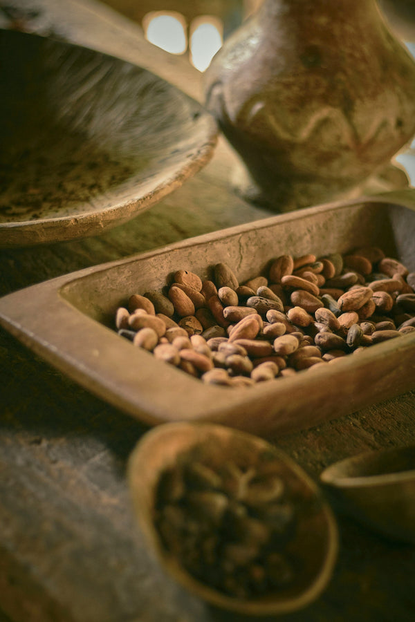 Cacao beans in a wooden tray with traditional processing tools, representing raw cacao sourcing and artisanal cacao preparation