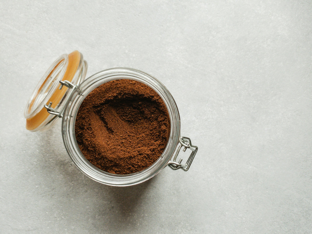 Open glass jar filled with coffee grounds on a light gray background