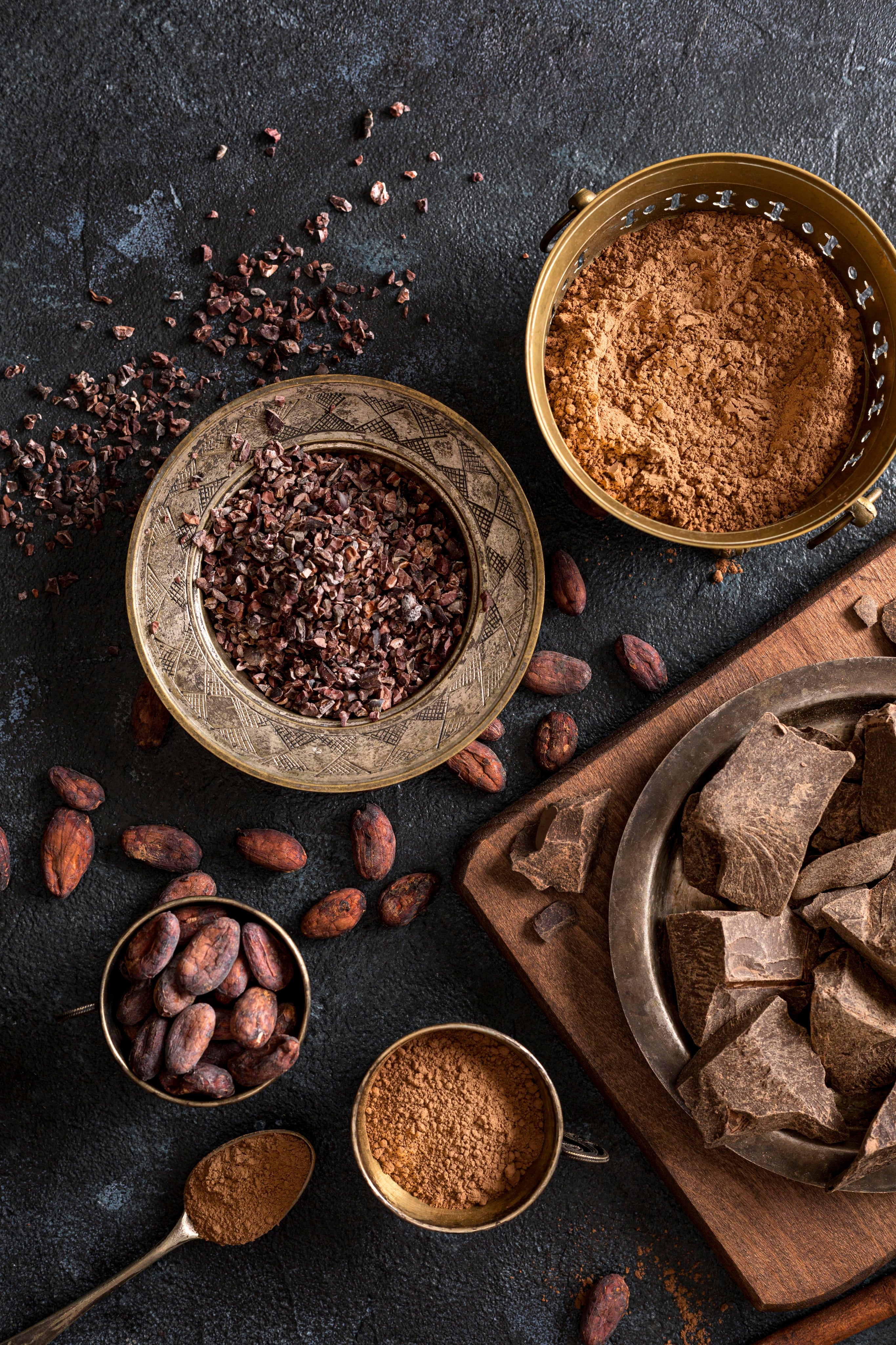 Cacao nibs, cacao powder, chocolate pieces, and cacao beans arranged on a dark surface, representing cacao processing and premium cacao product forms